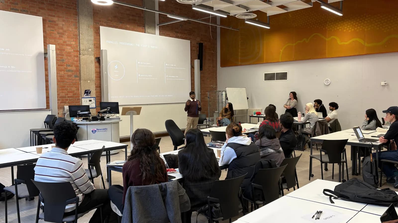 Tushar presenting to the programme in a Newcastle University seminar room, the cohort seated at tables in front of him, beside a slide titled Parallel processing: the first 6 months that breaks the first half year of an MSc into coursework, a part-time job, university roles and an internship